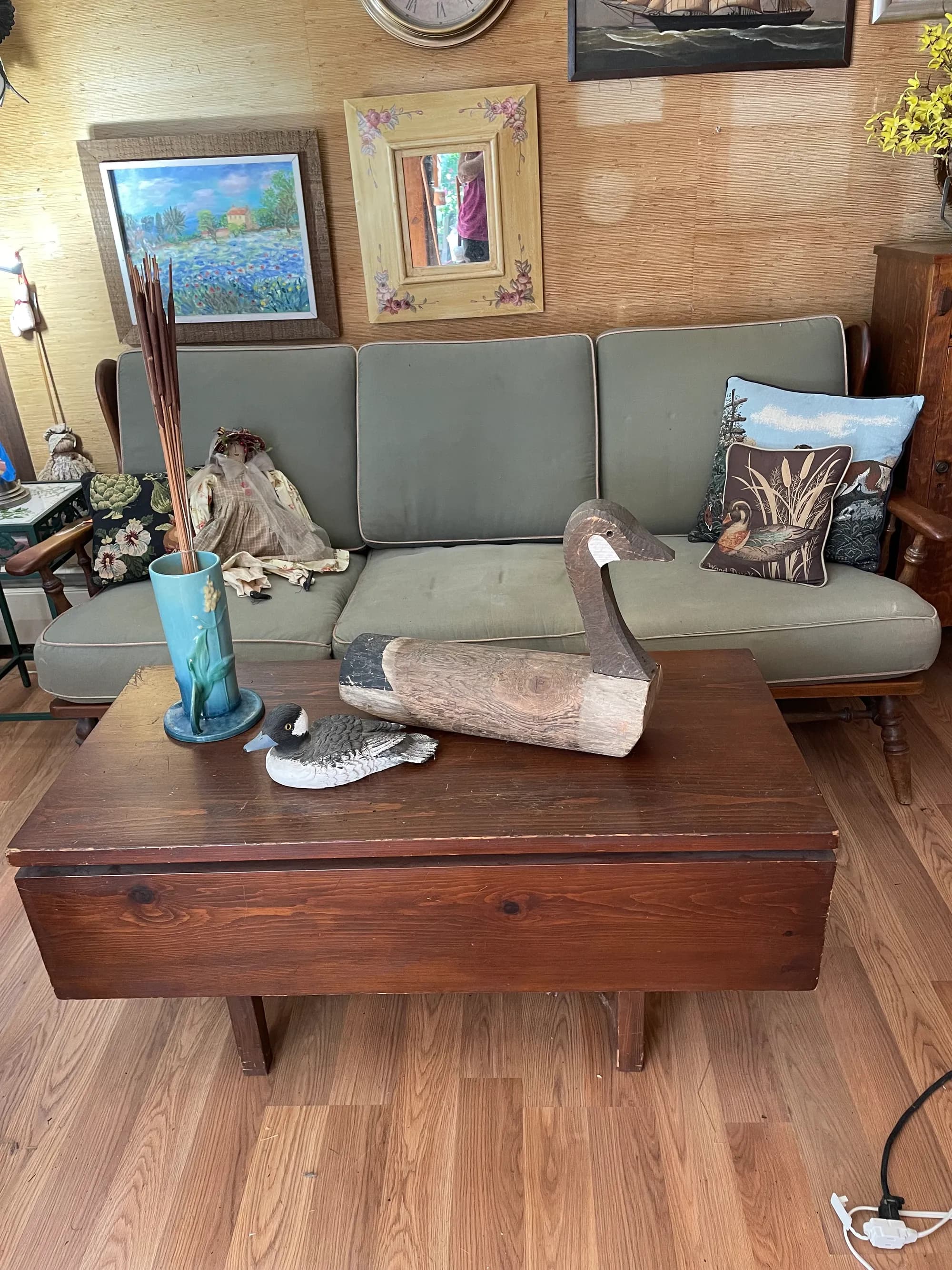 An estate-sale living room with a sage-green sofa, framed paintings, and carved wooden decoys.