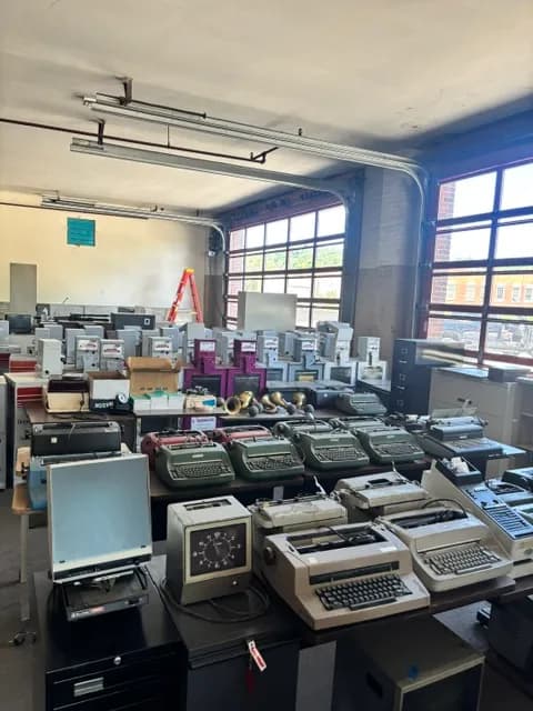 Rows of vintage typewriters and adding machines by tall windows.